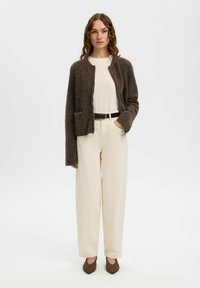 Femme aux cheveux ondulés portant un cardigan marron duveteux, une chemise crème, un pantalon crème large taille haute, une ceinture marron et des chaussures pointues marron, debout droite.