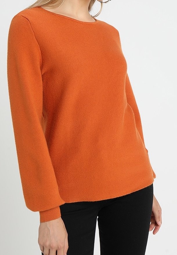 Femme portant un pull en laine orange à manches longues associé à un pantalon noir, debout devant un fond blanc uni.