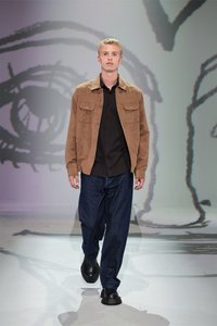 Jachetă din suede bej, cămașă neagră cu nasturi, pantaloni din denim albastru închis, de tip lejer, și pantofi negri masivi, purtată de un model masculin pe un fundal minimalist.