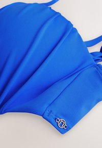 Haut de bikini bleu rembourré avec bretelles ajustables et un petit charm logo circulaire argenté sur le côté.