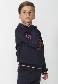 Sudadera navy con mangas de textura azul en contraste, puños y dobladillo acanalados, que presenta texto y acentos rojos en la parte frontal.