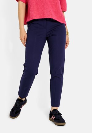 Mujer vistiendo pantalones ajustados azul marino, zapatillas negras con rayas rosas y un suéter rosa brillante y peludo, de pie frente a un fondo blanco.