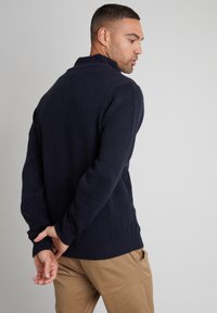 Granatowy sweter dziany z wysokim kołnierzem, teksturowanym materiałem i prążkowanym wykończeniem. Model ma ręce splecione za plecami, nosi beżowe spodnie.