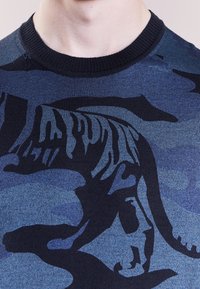 Pull en tricot bleu avec un design en silhouette de tigre noir. Encolure côtelée noire et tissu texturé. Gros plan mettant l'accent sur le motif et le contraste des couleurs.