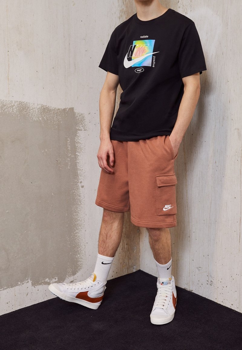 Hombre con camiseta negra de Nike con logo colorido, pantalones cortos cargo marrones de Nike, calcetines blancos de Nike y zapatillas blancas con detalles marrones.