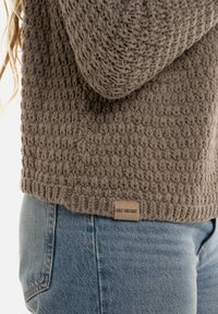 Bruine gebreide trui met een gestructureerd patroon, geribbelde zoom en een label van stof met de tekst "Knit Factory", gecombineerd met blauwe denimjeans.