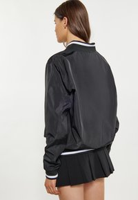Geacă de tip bomber neagră, confecționată din material lucios. Prezintă manșete ribbate și un guler cu dungi albe și bleumarin. Design clasic, cu o croială relaxată.