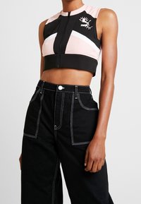 Top corto negro con diseño geométrico en rosa y blanco, con cierre frontal de cremallera y un logotipo. Combinado con pantalones negros de cintura alta con costuras blancas.