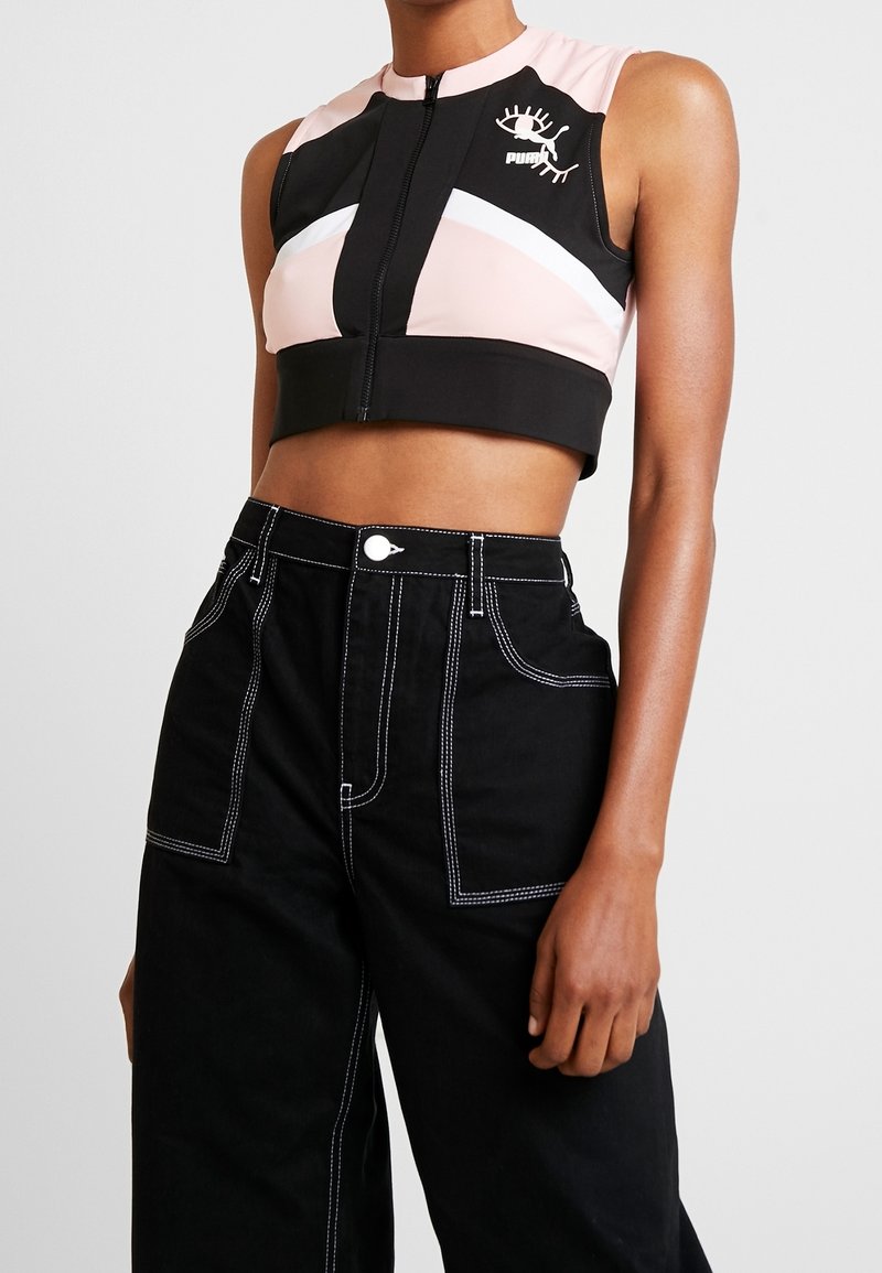 Top corto negro con diseño geométrico en rosa y blanco, con cierre frontal de cremallera y un logotipo. Combinado con pantalones negros de cintura alta con costuras blancas.