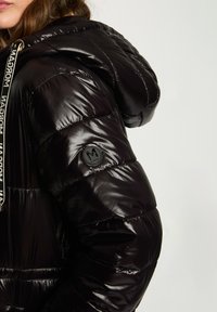 Chaqueta acolchada negra con una textura brillante y acolchada, que cuenta con capucha y un parche con logo en la manga. Detalle de cordón en el cuello.