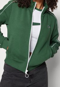 Vihreä vetoketjullinen huppari, joka on valmistettu pehmeästä kankaasta. Siinä on valkoiset saumat hihan varrella, korkea kaulus ja pieni logo rinnassa.