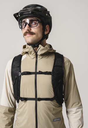 Mann mit Schnurrbart, der einen schwarzen Fahrradhelm, dunkle Schutzbrille, beige Jacke und schwarzen Rucksack mit Brustgurt trägt und leicht nach links schaut.