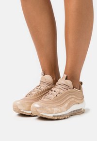 Šviesiai rudos Nike Air Max 97 sportbačiai su tekstūruotu paviršiumi, matoma „Air“ sistema ir švelniu blizgesiu; su sluoksnių dizainu ir apvaliais raišteliais.