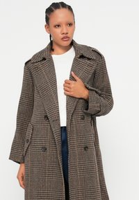 Double-breasted plaid coat in brown and red tones, featuring a notched collar, large buttons, and a textured fabric with a relaxed fit.