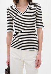 Frau trägt ein schwarz-weiß gestreiftes V-Ausschnitt-Shirt mit kurzen Ärmeln und weiße Hose, hält eine dunkelbraune Handtasche in ihrer linken Hand.