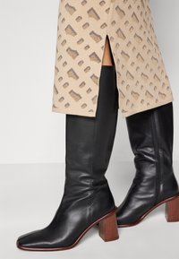 Stivali alti in pelle nera con punta quadrata e tacco in legno. Il design presenta una texture liscia e una silhouette aderente.
