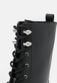 Gros plan d'une botte en cuir noir avec des lacets noirs passés à travers des œillets en argent et des crochets métalliques gravés du logo « MK ».