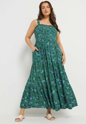 Frau in einem grünen, ärmellosen Maxikleid mit Blumenmuster, breiten Trägern und Sandalen, die mit einer Hand in der Hosentasche vor einem einfarbigen Hintergrund steht.