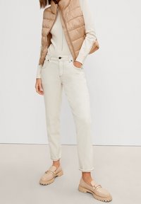 Béžová prošívaná vesta, krémový přiléhavý svetr, světlé džíny a béžové mokasíny s tlustou podrážkou. Hladká textura, ležérní design s kapsami.