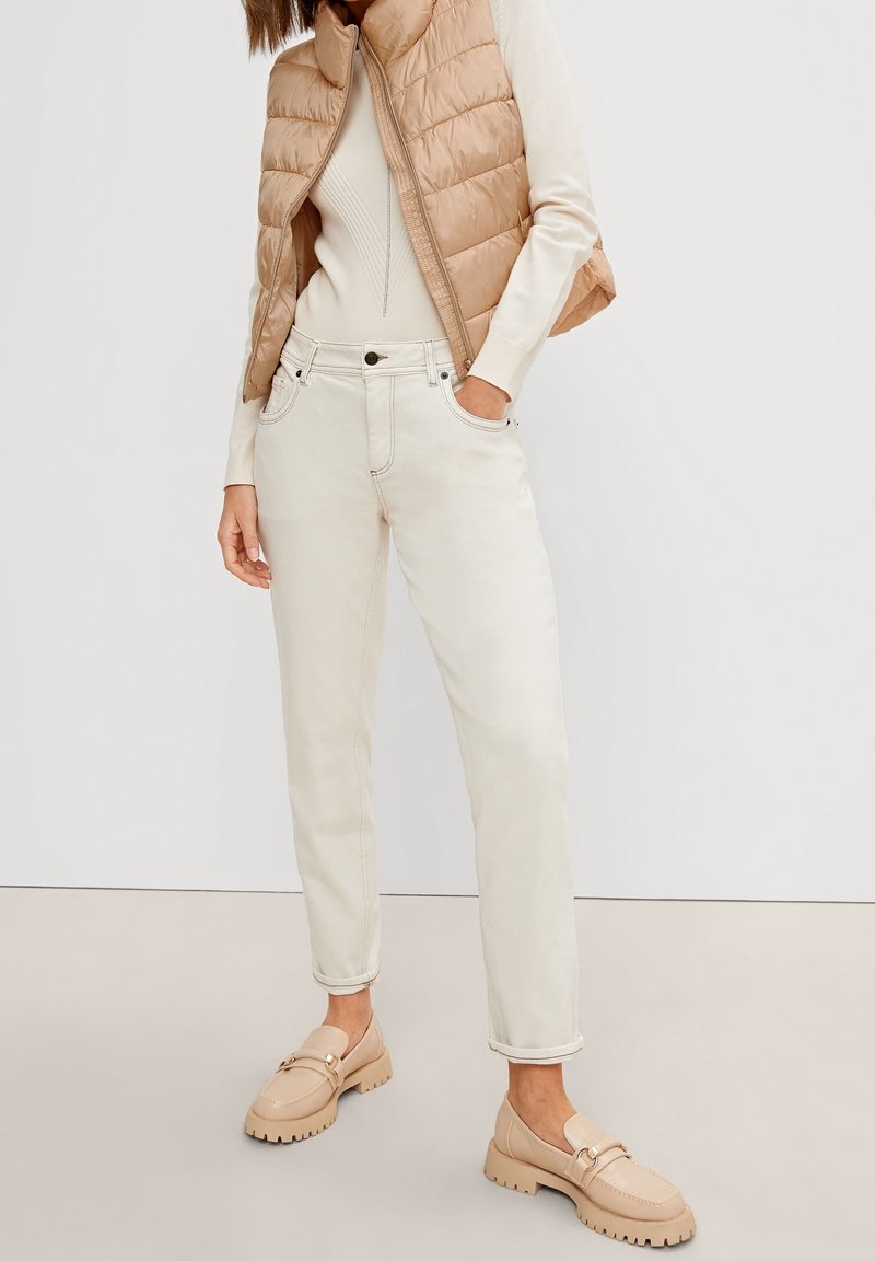 Béžová prošívaná vesta, krémový přiléhavý svetr, světlé džíny a béžové mokasíny s tlustou podrážkou. Hladká textura, ležérní design s kapsami.