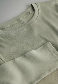 Gai zaļš ribots krekls ar garām piedurknēm, apaļu kakla izgriezumu un šuvēm, kuram ir maiga tekstūra un viegla spīduma pakāpe.