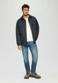 Navy katoenen shirtjas met twee borstzakken, gecombineerd met een wit shirt, blauwe jeans en bruine veterschoenen. Eenvoudig en functioneel ontwerp.