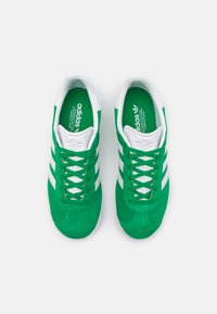 Green suede sneakers with white rubber soles, featuring three white stripes on the sides and green laces, showcasing a minimalist design.