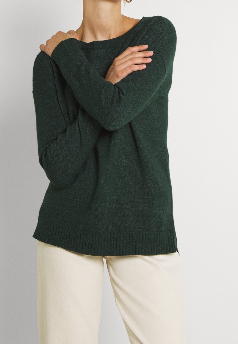 Dunkelgrüner Strickpullover mit lockerer Passform, gerippten Bündchen und Saum sowie weitem Ausschnitt. Kombiniert mit hellen, geraden Hosen.