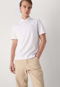 Polo branco de manga curta com colarinho, três botões e um pequeno logótipo no peito, combinado com calças leves em bege.