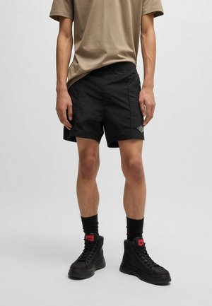 Hombre con camiseta beige, pantalones cortos negros con logo en la pierna izquierda, calcetines negros hasta la mitad de la pantorrilla y zapatillas negras de caña alta con detalles en rojo.