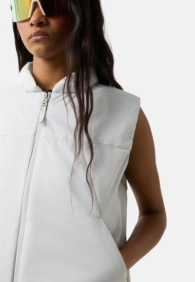 Młoda kobieta w białej kamizelce na suwak bez rękawów i futurystycznych dużych lustrzanych okularach przeciwsłonecznych na jednolitym tle.