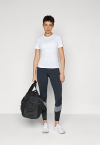 Hvit kortermet treningstrøye kombinert med marineblå tights med grå detaljer, og en svart treningsbag holdt i den ene hånden. Hvite sneakers.