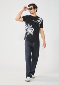 T-shirt noir à manches courtes avec un graphique de palmier blanc, associé à un jean bleu foncé et des baskets blanches. Un homme ajuste ses lunettes de soleil en marchant.