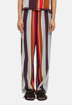 Osoba nosząca luźne, wielokolorowe spodnie w pionowe paski, pasującą koszulę i czarne sandały z frędzlami, stojąca na białym tle.