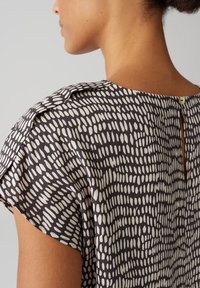 Korte mouwen blouse van donkergrijze stof met een onregelmatig wit stippenpatroon. Heeft een keyhole terugdetail en een zachte textuur.