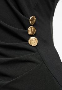 Juodos suknelės audinys pasižymi rietu tekstūra su trimis metaliniais, netaisyklingos formos auksiniais mygtukais kaip dekoratyviniais akcentais.