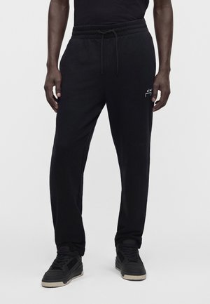 Homme portant un pantalon de jogging noir avec taille à cordon et des baskets noires, debout devant un fond blanc uni.