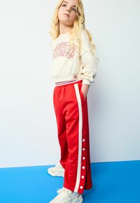 Røde sweatpants med hvide sidelommer og trykknapper, parret med en kremfarvet sweatshirt med teksten "HAPPINESS" i pink. Teksturstof.