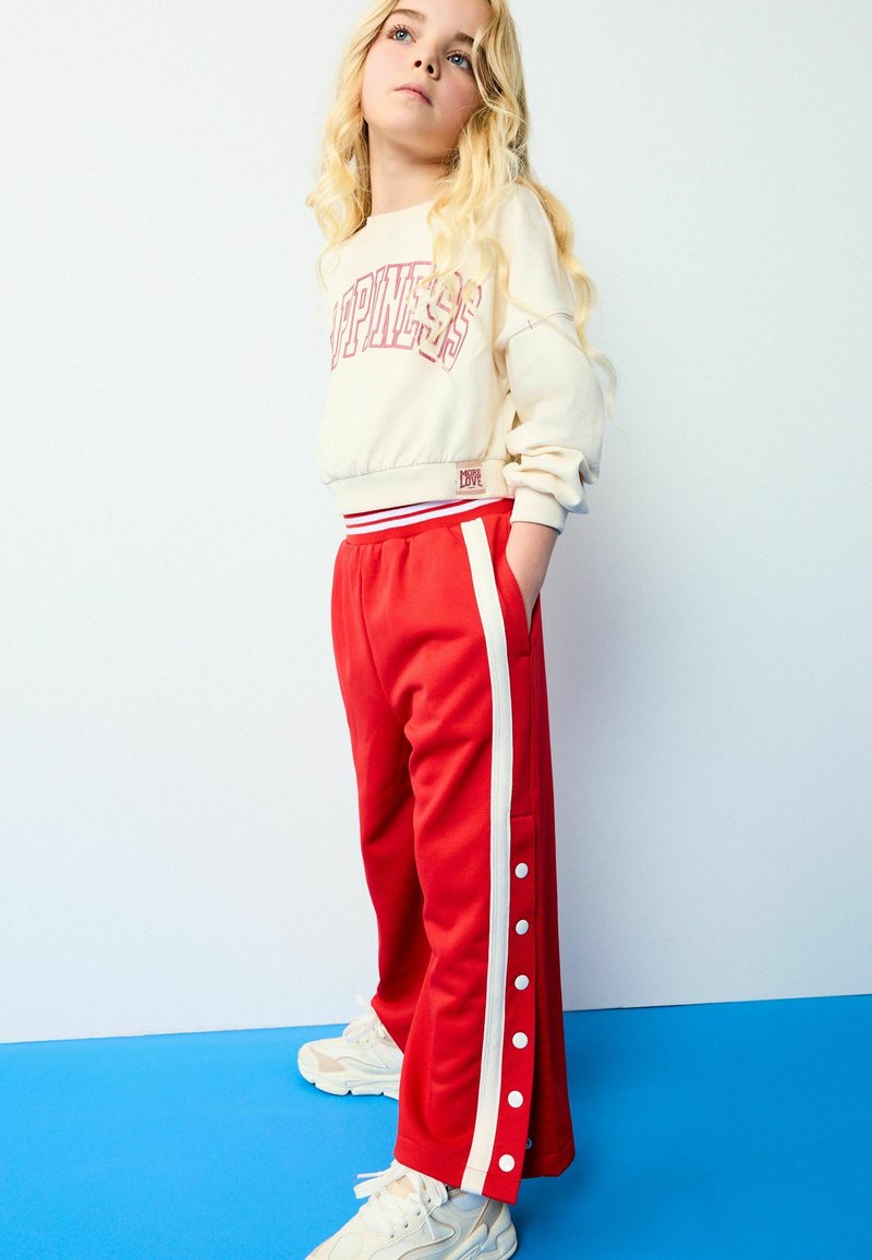 Røde sweatpants med hvide sidelommer og trykknapper, parret med en kremfarvet sweatshirt med teksten "HAPPINESS" i pink. Teksturstof.