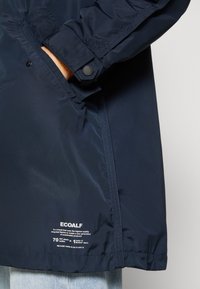 Détail de la manche et de la poche latérale d'une veste navy avec une main à l'intérieur, logo blanc ECOALF et texte sur la durabilité près de l'ourlet, portée sur un jean bleu clair.
