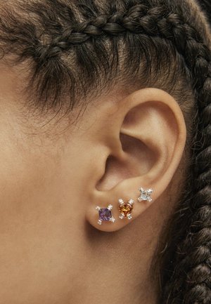 Tres pendientes exhibidos en una oreja; con piedras preciosas moradas, naranjas y transparentes engastadas en metal de tono plateado con configuraciones decorativas de garras.