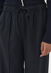 Persoon in zwarte losse broek met elastische tailleband en gestrikte koord, en een donker geribbeld langemouwen top, hand ontspannen naast zich.