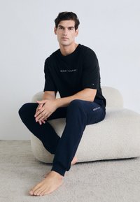 Camiseta de algodón negra con el logo blanco de "TOMMY HILFIGER", combinada con pantalones de chándal azul marino. Textura suave, corte relajado y sin patrones visibles.