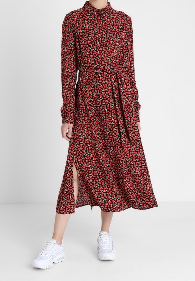 Femme portant une robe midi rouge à fleurs à manches longues avec une fente latérale et des baskets blanches, debout devant un fond blanc uni.