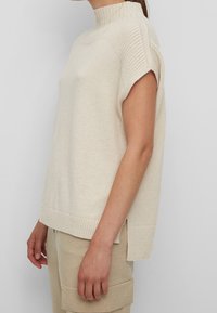 Pull en crème à manches courtes avec un col haut, des accents côtelés sur les épaules et des fentes sur les côtés pour un détail supplémentaire. Texture douce.