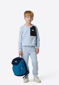 Sudadera y pantalones azul claro con detalle de bolsillo negro, con un logo. El niño lleva una mochila azul con solapa y detalles de fieltro.