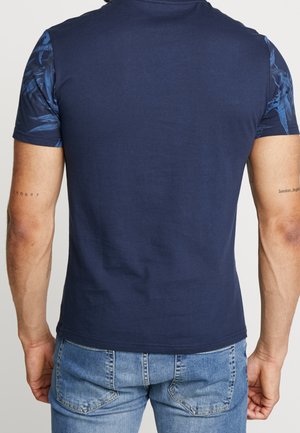 Homme portant un t-shirt bleu marine avec des manches courtes à motifs bleu foncé et un jean bleu clair, vu de dos.