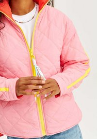 Prošívaná růžová bunda se žlutým zipem a akcenty. Obsahuje oranžovou kapuci a vícedimenzionální táhlo zipu. Ruce umístěné kolem zipu.