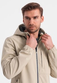 Beige hoodie-jacka med elastiska ärmslut, dragkedja fram och bröstficka. Slät, vattenavvisande tyg med en texturerad innerfoder.