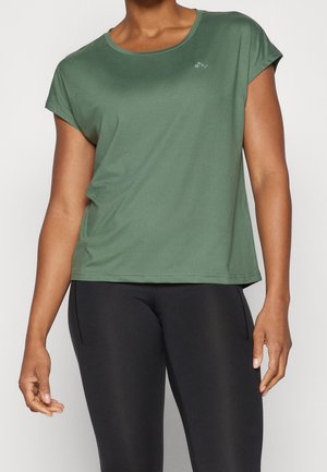 Person trägt ein olivgrünes kurzärmeliges Sportshirt und schwarze enganliegende Leggings, steht vor neutralem Hintergrund.